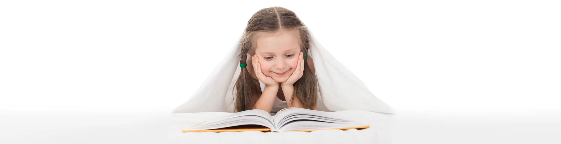 A young girl lying under a blanket, smiling as she reads an open book